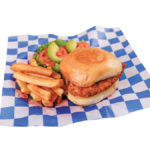 Crispy chicken sandwich, sliced tomatoes, avocado slices, and french fries on blue and white checkered paper