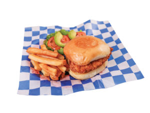 Crispy chicken sandwich, sliced tomatoes, avocado slices, and french fries on blue and white checkered paper