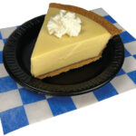 Slice of Key Lime pie with a dollop of whipped cream. On a small black plate placed on a white and blue checkered paper